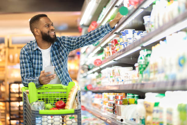 Black Male Buyer Shopping Groceries In Supermarket Taking Dairy Product From Shelf Standing With Shop Cart Indoors.